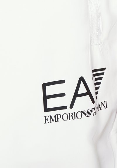 Бяла тъкан, показваща голям черен лого с надпис "EA7 EMPORIO ARMANI" с хоризонтални ивици, подчертаваща модерен дизайн и брандиране.
