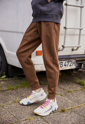 Osoba w brązowych spodniach dresowych Nike i białych trampkach z różowymi sznurowadłami stoi na brukowanej powierzchni w pobliżu białego pojazdu.