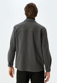 Grå skjorte med knapper set fra ryggen, der har en struktureret tekstur, buet kant og opsmøgede ærmer med knapper. Enkel krave design.