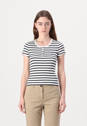 Csíkos, rövid ujjú felső fehér és sötétkék színben, kerek nyakkivágással, gombos záródással és bordás textúrával. Bézs nadrággal viselve.