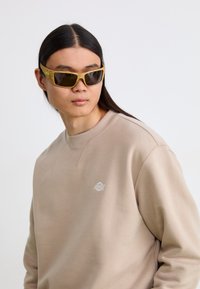 Occhiali da sole con una montatura spessa di colore marrone chiaro e lenti scure, indossati con una felpa beige con un piccolo logo sul petto.