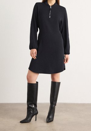 Femme portant une robe noire longue à manches longues, longueur genou, avec une fermeture éclair à l'avant et des bottes noires à talons hauts jusqu'au genou sur un fond uni.