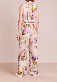 Bloemige jumpsuit in pastelpink. Mouwloos ontwerp met een hoge hals, brede broekspijpen en levendige, multicolore bloemenpatronen. Witte schoenen.