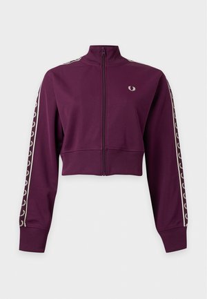 Femme portant une veste courte bordeaux zippée avec des manches à motifs, une jupe midi plissée noire, des chaussettes grises et des mocassins noirs, debout sur fond blanc.