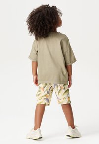 Olive green oversized koszula z krótkim rękawem, wzorzyste kremowe szorty z ananasami i liśćmi, noszone z białymi tenisówkami. Naturalne kręcone włosy.