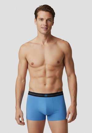 Homme torse nu debout, portant un boxer bleu avec une ceinture noire, sur un fond clair uni.
