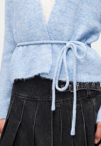 Ανοιχτό μπλε χνουδωτό ζακέτα δεμένη με ζώνη πάνω από λευκό τοπ, συνδυασμένη με μαύρη πλισέ φούστα με ψηλή μέση και ζώνη με ασημένια αγκράφα.