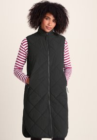 Woman with curly hair wearing a long black quilted vest over a white and pink striped long-sleeve shirt, hands in vest pockets.