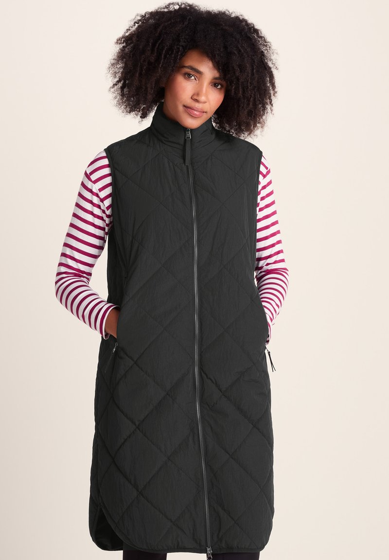 Woman with curly hair wearing a long black quilted vest over a white and pink striped long-sleeve shirt, hands in vest pockets.