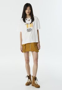 T-shirt graphique blanc avec texte noir et motif coloré au-dessus ; associé à une jupe moutarde avec une fermeture éclair à l'avant et des poches ; bottes en cuir lacées marron.