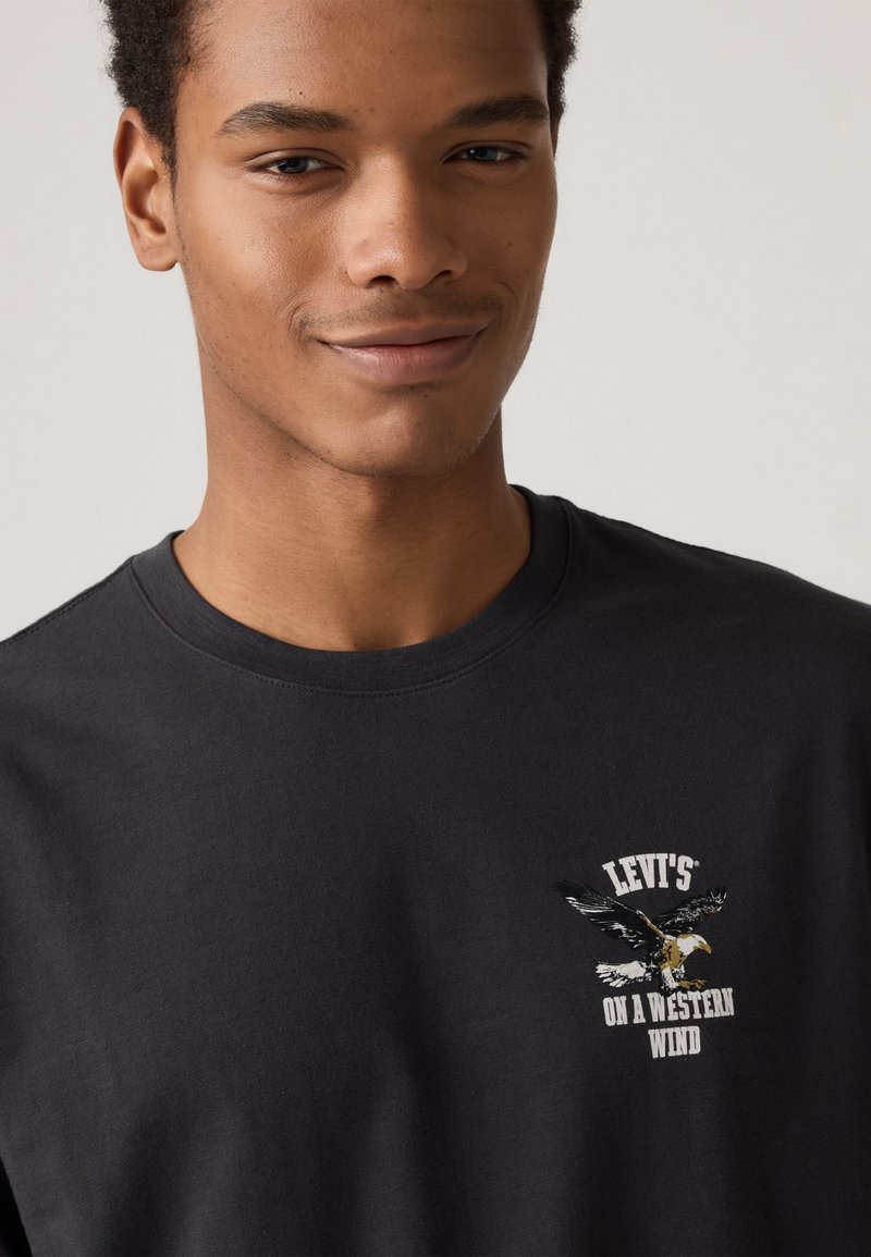 T-shirt en coton noir avec un imprimé graphique d'un aigle et le texte "LEVI'S ON A WESTERN WIND" en blanc et jaune. Design classique à encolure ronde.