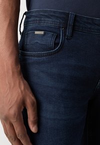 Gros plan d’une main posée à côté de la poche avant d’un jean en denim bleu foncé avec une petite étiquette de marque en métal.