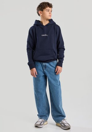 Dunkelblaue Kapuzenjacke mit einer Fronttasche und gesticktem Text, kombiniert mit hellblauen weiten Jeans und grauen Sneakers mit schwarzen Akzenten.