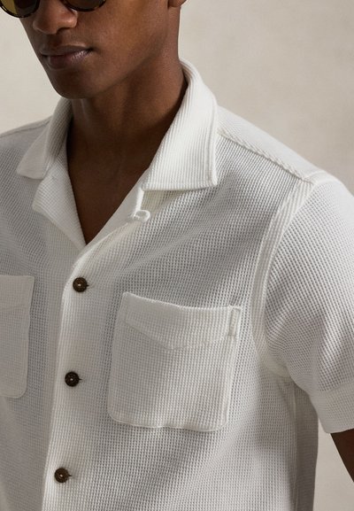 Λευκό πουκάμισο κοντό μανίκι σε υφή πλεκτού με μυτερό γιακά, δύο εμπρόσθιες τσέπες και καφέ κουμπιά. Χαλαρή εφαρμογή και ελαφρύ ύφασμα.