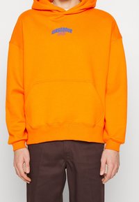 Sudadera con capucha naranja y bolsillo frontal, con texto bordado azul en el pecho. La tela parece suave y texturizada.
