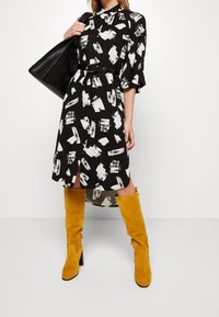 Abito nero con motivo astratto bianco, maniche corte, vita con cintura e spacco laterale. Abbinato a stivali gialli in suede al ginocchio e una borsa nera.
