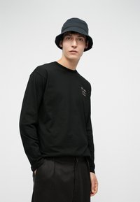 Tânăr îmbrăcat cu o pălărie de tip bucket neagră, cu o bluză lungă neagră cu text japonez pe piept și pantaloni negri, stând în fața unui fundal deschis.