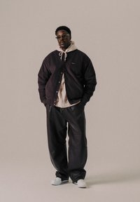 Black cord jacket over a light brown hoodie, paired with black leather pants and white slip-on shoes, on a neutral background.