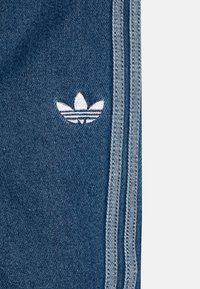 Denim tkanina v temno modri barvi z belim vezenim logotipom in sivimi stranskimi črtami. Ima gladko teksturo in tipičen atletski kroj.