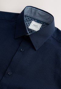Marineblå skjorte med et tekstureret mønster, der har en spids krave og en kontrasterende foring med geometrisk print. Inkluderer knaplukning.