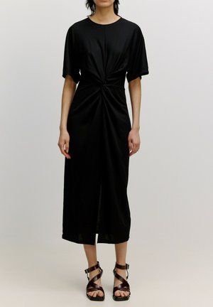 Schwarzes Midi-Kleid mit verdrehtem Vorderteil, weichem Stoff, kurzen Ärmeln und einem mittigen Schlitz. Kombiniert mit braunen Riemchensandalen.