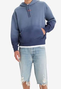 Felpa con cappuccio in gradiente blu, con tasca frontale e logo rosso. Abbinata a pantaloni shorts in denim strappati, con tonalità blu predominanti.