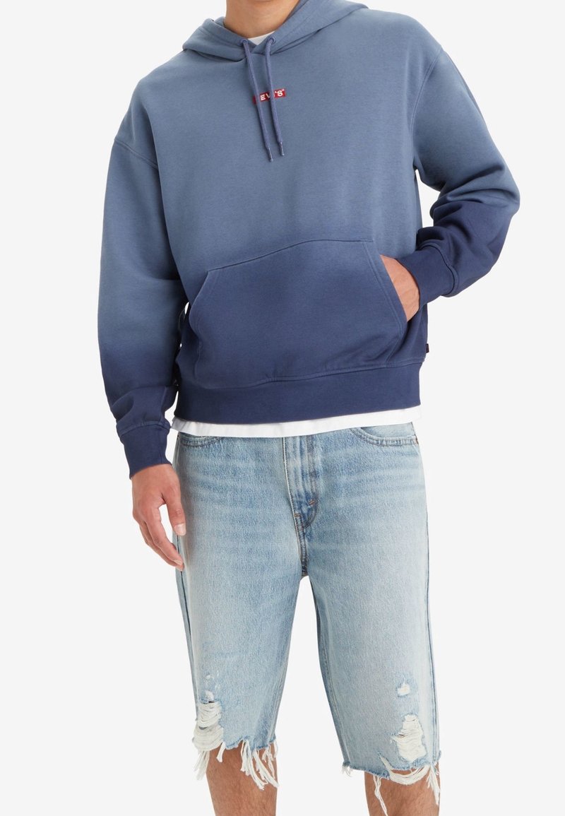Felpa con cappuccio in gradiente blu, con tasca frontale e logo rosso. Abbinata a pantaloni shorts in denim strappati, con tonalità blu predominanti.