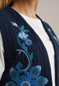 Laivastonsininen tikattu liivi, jossa on sinistä kukkakirjontaa eri sävyissä, monimutkaisia yksityiskohtia ja teksturoidun kankaan pinta.