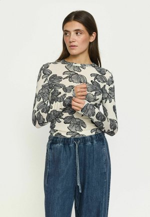 Femme aux longs cheveux bruns porte un haut beige à manches longues avec un imprimé floral noir et un pantalon en jean bleu ample, regardant vers sa gauche.