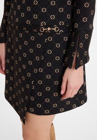 Nahaufnahme eines schwarzen Kleides mit beige geometrischen Mustern, einem goldenen Schnallendetail in der Taille und einem Schlitz am Ärmel.