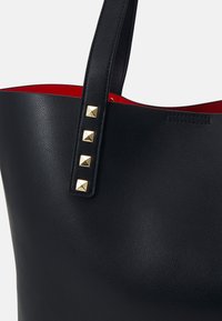 Tote en cuir noir avec une texture lisse, présentant un intérieur rouge et une sangle ornée de quatre clous en forme de pyramide dorés.