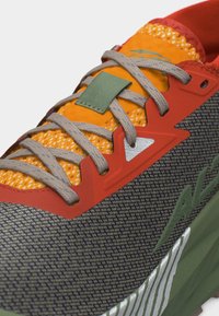 Κοντινή λήψη ενός πολύχρωμου αθλητικού παπουτσιού με πορτοκαλί δίχτυ, κόκκινες επενδύσεις, γκρι κορδόνια, πράσινη θηλιά έλξης και υφασμένη πράσινη σόλα.