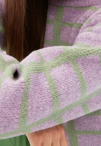 Sweter dziergany w jasnofioletowym kolorze z zielonym wzorem w kratę. Materiał wydaje się miękki i ma fakturowane wykończenie. Posiada szeroki kołnierz i dopasowane rękawy.