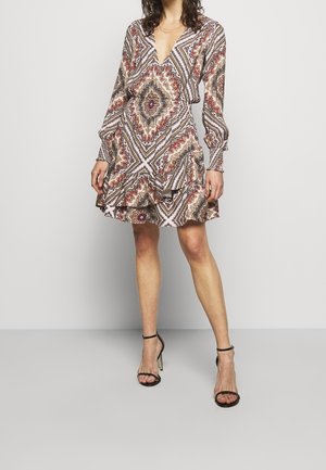 Nainen pukeutuneena kuviolliseen pitkähihaseen kietaisumekkoon, jossa on röyhelöhelma, ja mustiin nilkkaremmiin kiinnitettyihin korkeakorkoisiin sandaaleihin, seisomassa yksiväristä taustaa vasten.