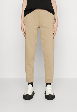 Beige taps toelopende joggingbroek gedragen met zwarte sokken en witte plateau sneakers, gecombineerd met een zwart shirt, staand op een grijze vloer.