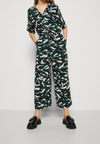 Combinaison raccourcie en vert foncé avec des motifs abstraits blancs et noirs, dotée d'un col, de manches courtes et de poches latérales. Portée avec des chaussures à plateforme.