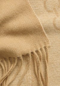 Sciarpa in lana beige con una trama morbida, caratterizzata da bordi frangiati e un motivo sottile, delicatamente appoggiata per mostrare la profondità del tessuto e le variazioni di colore.