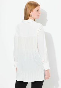 Blusa bianca con retro plissettato, maniche lunghe e polsini con bottoni. Il tessuto liscio e il colletto aggiungono struttura al design.