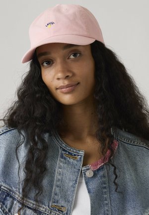 Młoda kobieta z długimi kręconymi włosami, w różowej czapce Levi’s i dżinsowej kurtce na białym topie, neutralne tło.