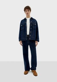 Denim jacket and pants in dark wash with brown stitching; white t-shirt underneath; brown shoes; relaxed fit design; four-button closure.