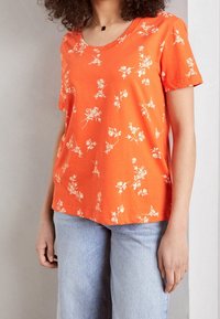 Orange kortærmet t-shirt med et hvidt blomstermønster, rund halsudskæring og en afslappet pasform, parret med lyseblå denimjeans.