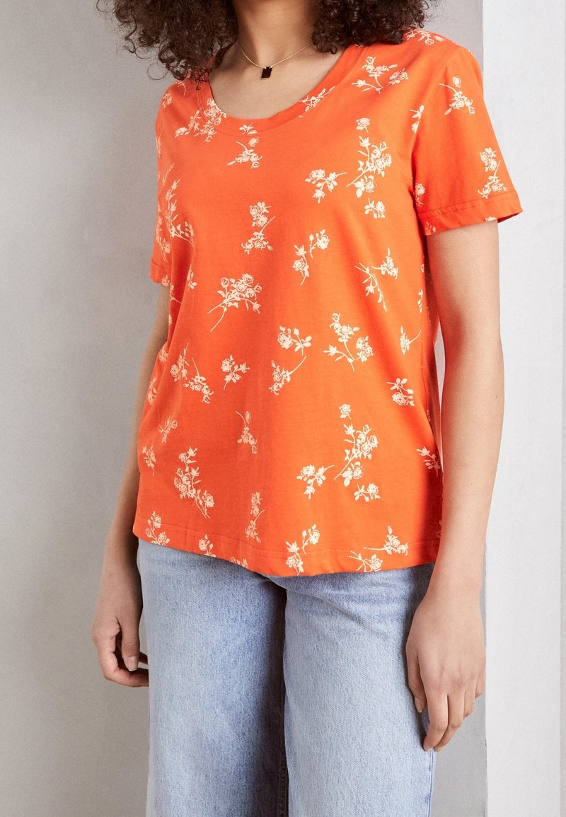 Orange kortærmet t-shirt med et hvidt blomstermønster, rund halsudskæring og en afslappet pasform, parret med lyseblå denimjeans.