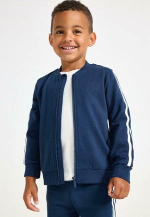 Donkerblauwe ritsjack met witte zijstrepen, ribgebreide manchetten en kraag. Draag over een wit t-shirt. Gladde stof, getailleerde pasvorm.
