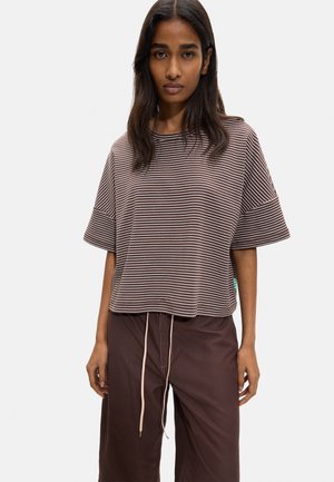 Kobieta z długimi ciemnymi włosami, w brązowo-białej pasiastym krótkim rękawie i ciemnobrązowych spodniach z jasnymi sznurkami.