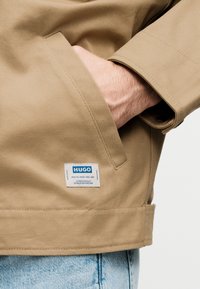 Τσέπη μπεζ υφασμάτινου σακακιού με ένα χέρι εν μέρει μέσα και μια λευκή ετικέτα με την επιγραφή "HUGO" πάνω από ανοιχτόχρωμο τζιν παντελόνι.