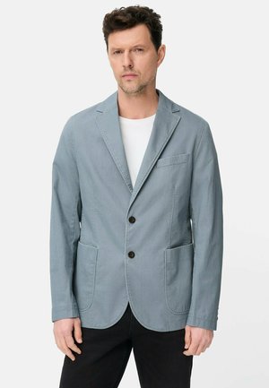 Mężczyzna noszący jasnoniebieską casualową marynarkę na białej koszuli z czarnymi spodniami, stojący na białym tle.