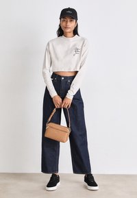 Gekürztes cremefarbenes Langarmshirt mit schwarzem Logo, hoch taillierte, dunkelblaue, weite Jeans, schwarze Sneakers und eine kleine beige Handtasche.