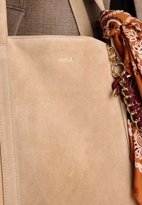 Beige suedine tote bag met een verguld logo, waaraan een bruine sjaal met patroon is geknoopt aan een vergulde ring.