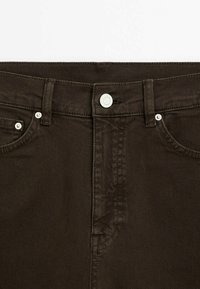 Gros plan sur un jean en denim marron foncé montrant le bouton, les passants de ceinture, les poches avant et les détails de couture.