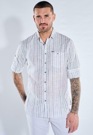 Mann mit kurzen Haaren und Tätowierungen trägt hellgraues gestreiftes Hemd mit Knöpfen und aufgekrempelten Ärmeln sowie weiße Hose, steht vor einfachem Hintergrund.